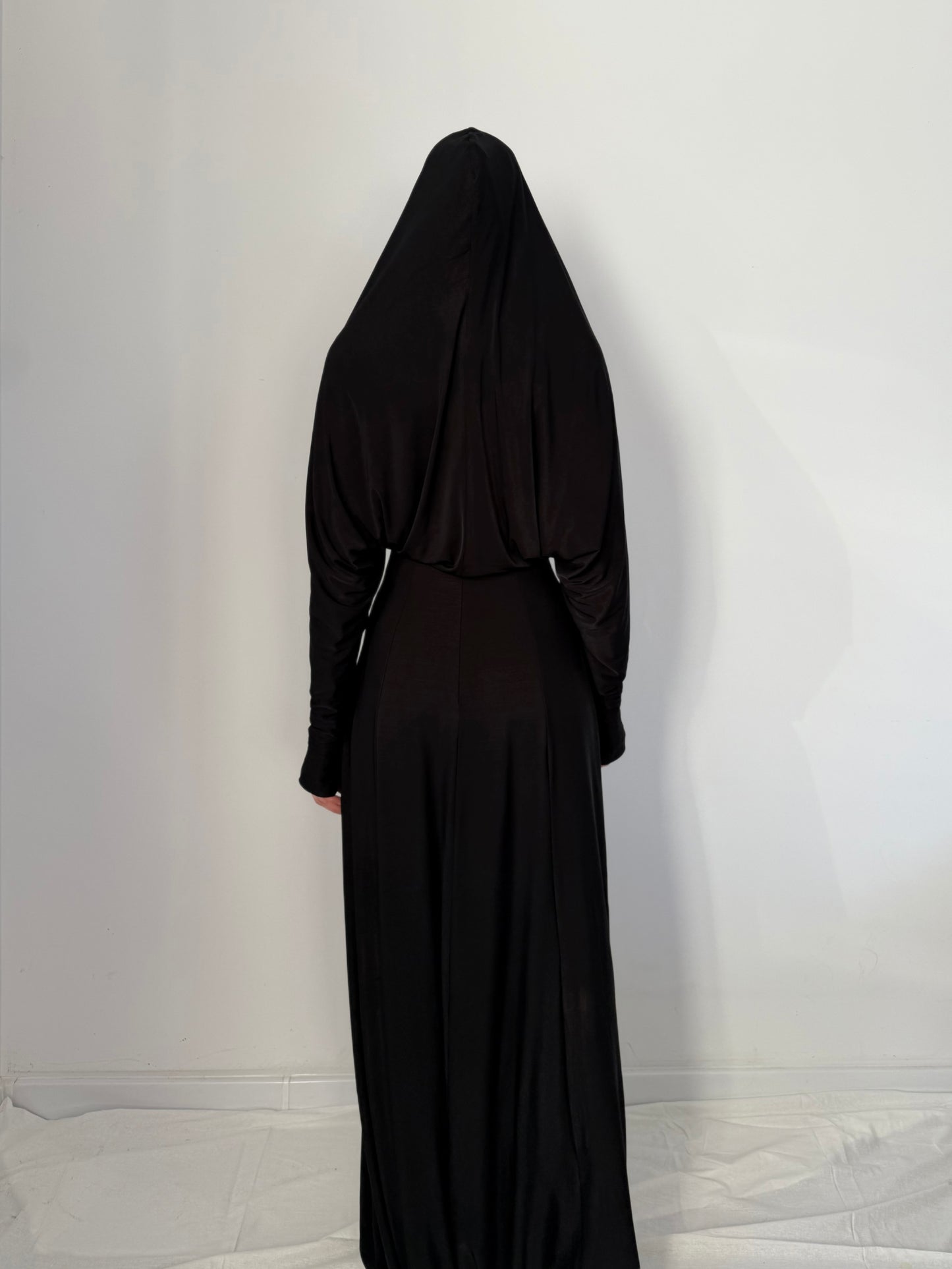 Person wearing a black dress with hood against a white wall