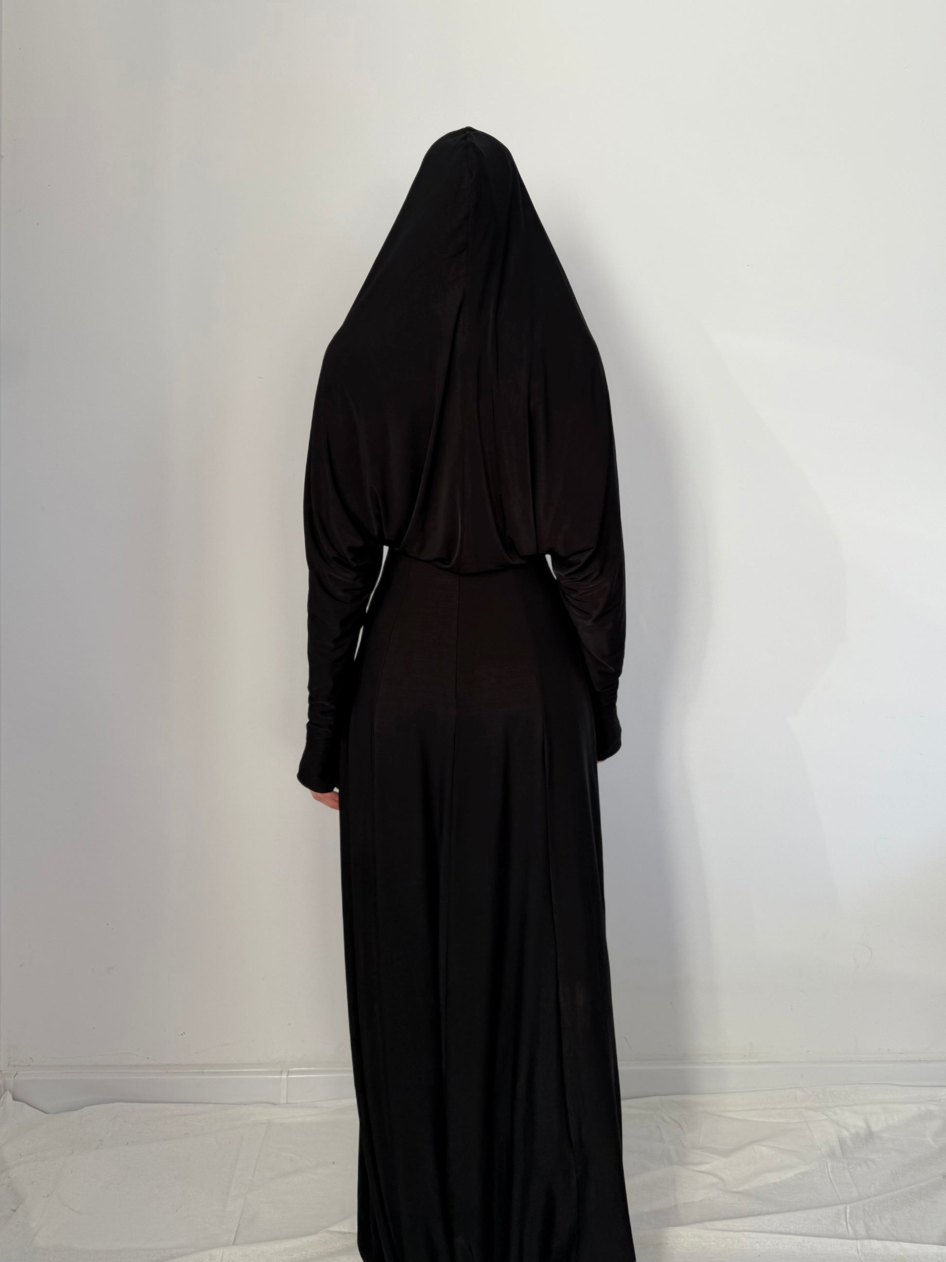 Person wearing a black dress with hood against a white wall